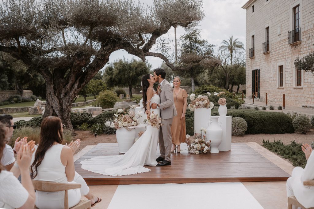 Bride and groom sharing a kiss during an elegant outdoor wedding ceremony in a historic Mallorca estate, surrounded by guests and floral décor.