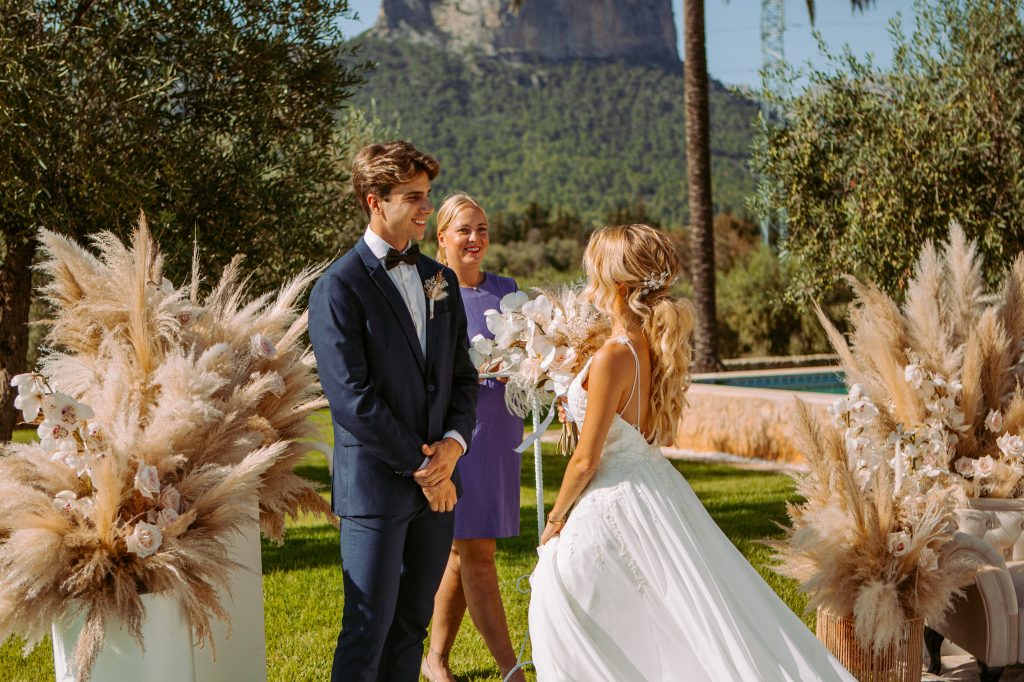 Outdoor wedding ceremony in Mallorca with the bride and groom standing beneath pampas grass arrangements, surrounded by olive trees and mountain views.