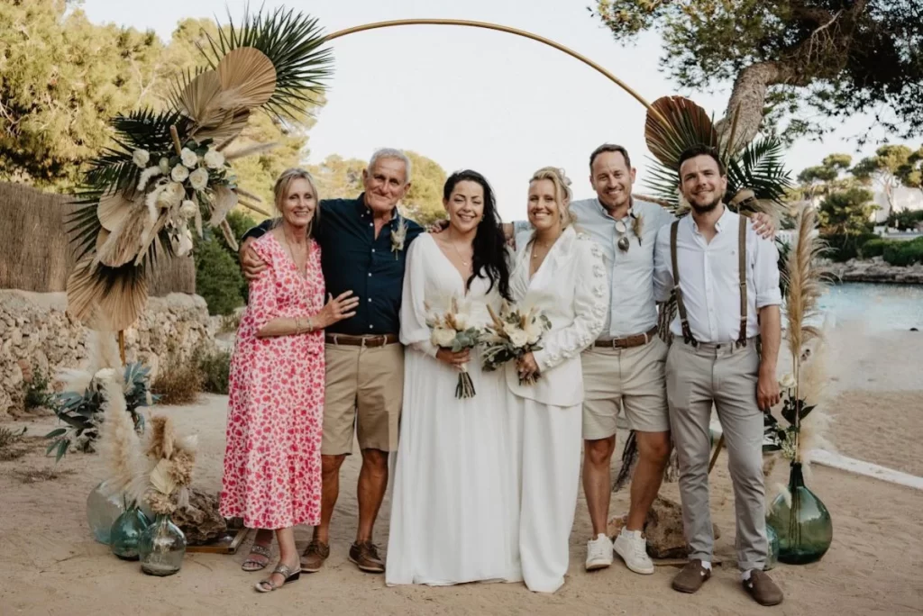 Two brides posing with their closest family and friends during an intimate same-sex beach wedding ceremony in Mallorca.